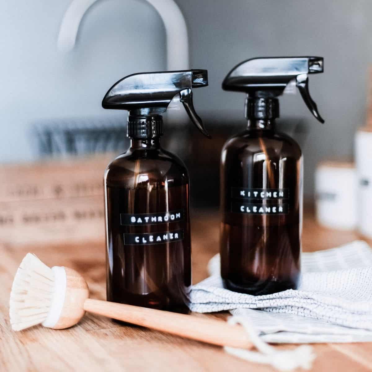 Two amber glass spray bottles labeled Bathroom Cleaner and Kitchen Cleaner sit on a wooden countertop, ready for action. A wooden dish brush and striped cloth lie nearby, complementing your cleaning schedule. In the background, a blurred kitchen sink hints at tasks yet to come.
