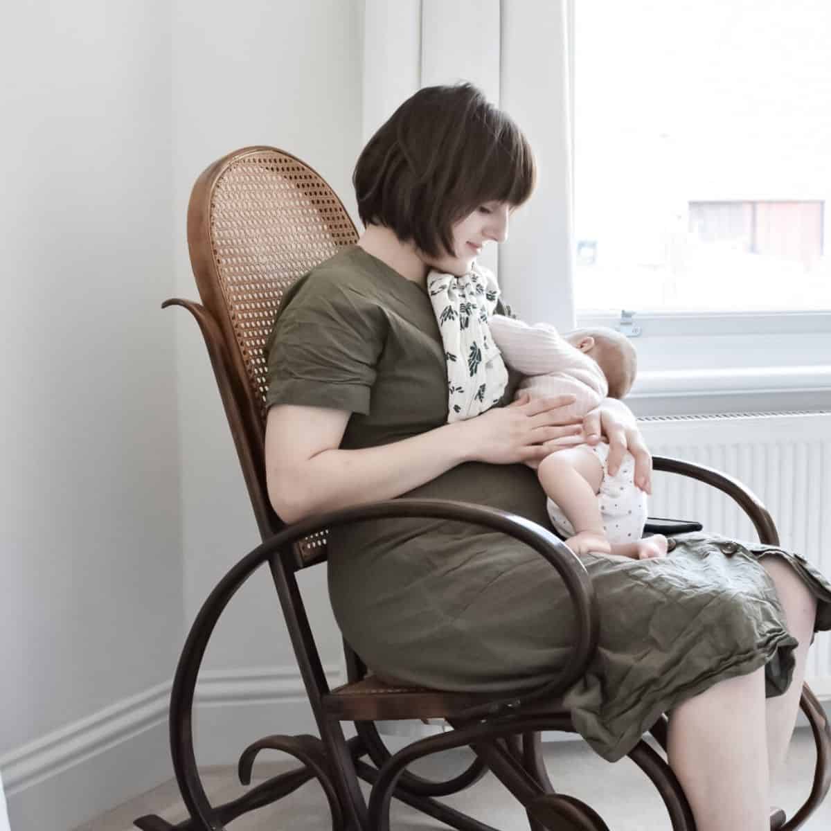 A woman sits in a wooden rocking chair by a window, tenderly breastfeeding her baby wrapped in a blanket. She gazes at the little one, who rests comfortably on her chest. The room is softly lit, with white walls and light-colored carpet, setting the scene of this gentle story.