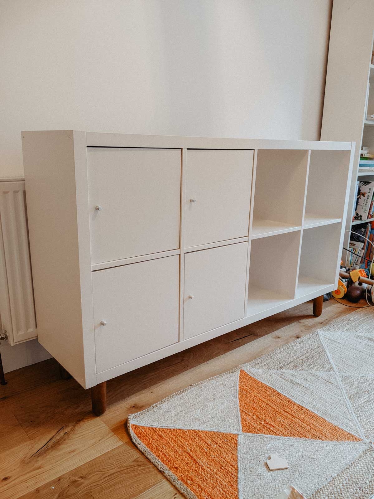 A white, six-cube storage unit with three open shelves and three cube doors, styled as an Ikea Kallax hack, stands on wooden legs next to a radiator. A geometric-patterned rug and part of a bookshelf are visible nearby.