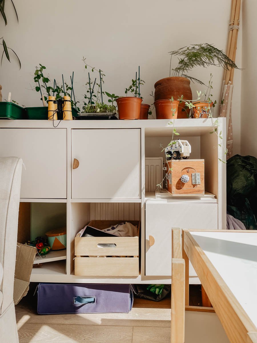 A pale pink Kallax unit displays potted plants, a wooden crate, a toy car, and small objects in sunlight. A beige chair and a wooden table are partially visible in the foreground.