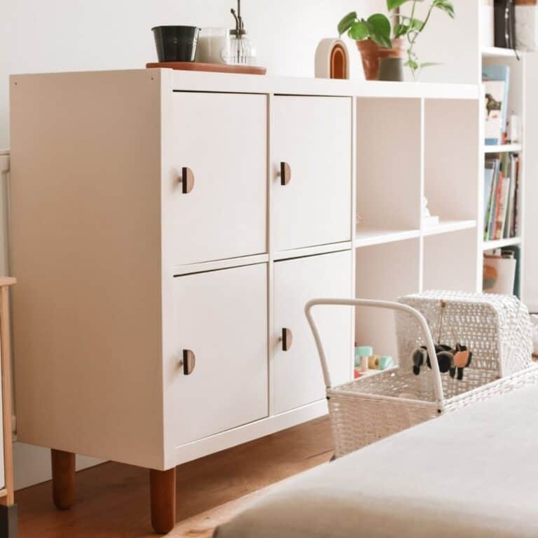 A white cabinet with wooden legs, resembling an IKEA Kallax hack, stands in a bright room. It features four closed compartments with circular handles and open shelves displaying small items. Nearby sits a wicker basket, while plants and books adorn the top and sides of the cabinet.