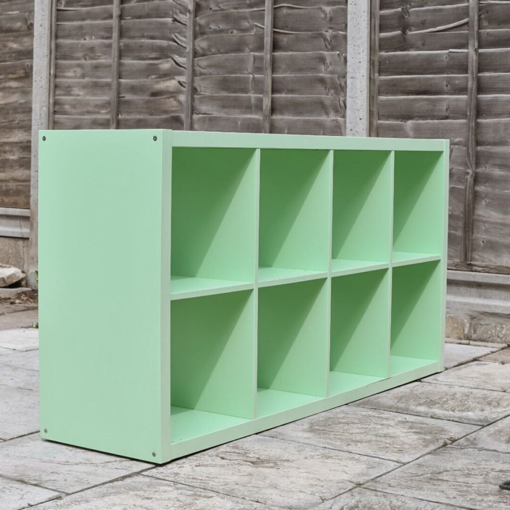 A light green, rectangular, eight-cube storage shelf sits on a stone patio in front of a weathered wooden fence.