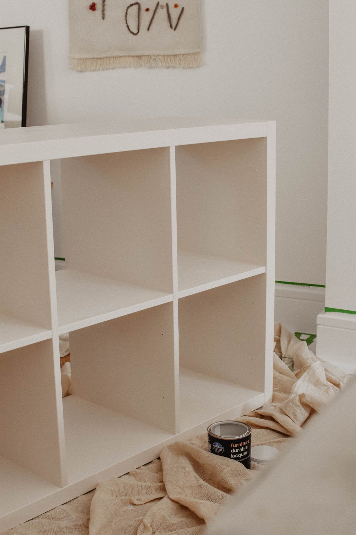 A pale pink IKEA cubby shelf sits on a canvas drop cloth next to a can of furniture lacquer in a room being redecorated. A framed picture and wall hanging are partially visible in the background.