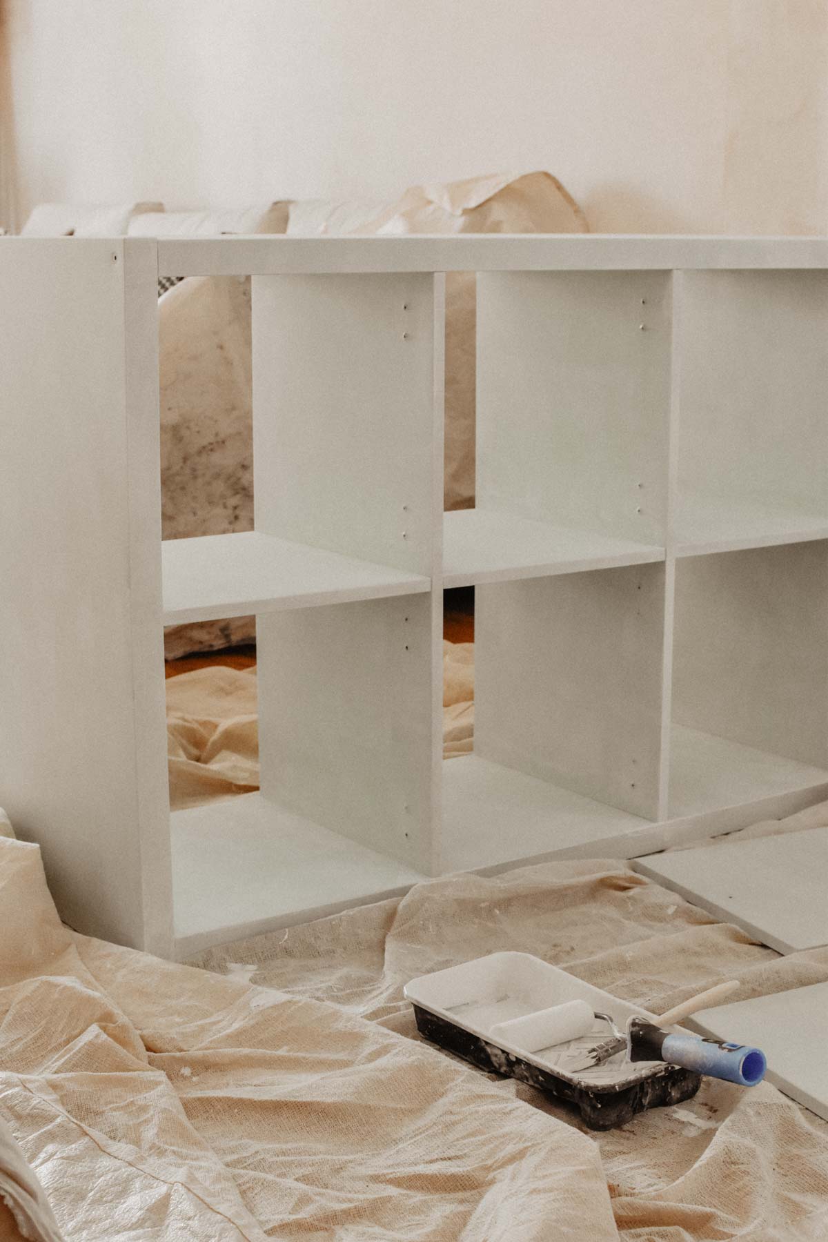 A white wooden cubby Ikea Kallax shelf is being painted. Drop cloths cover the floor, and painting supplies, including a roller and tray, are nearby in this bright, well-lit room.
