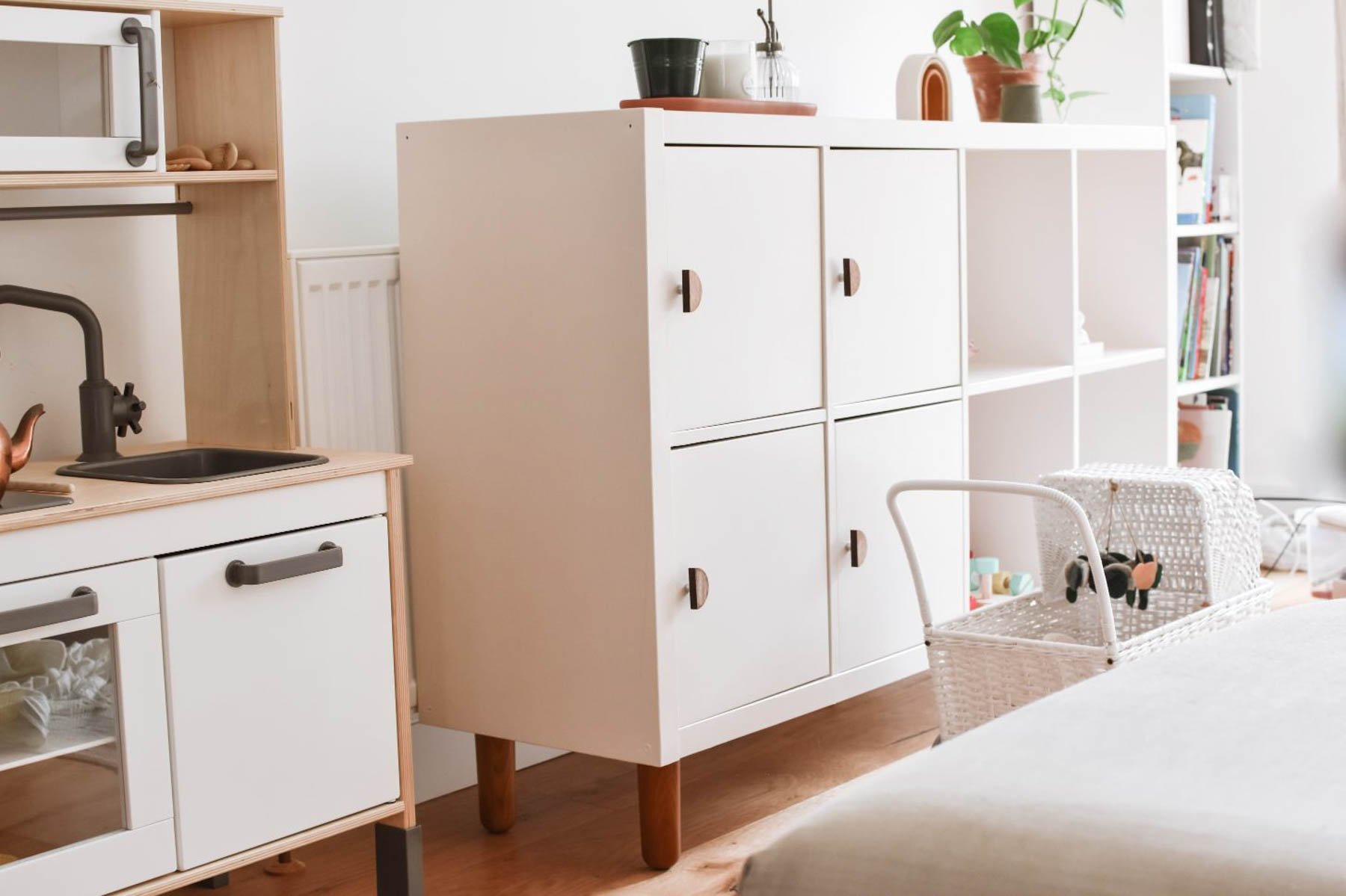 A tidy, modern living space featuring white cabinets with wooden handles,  a kitchen playset on the left, woven baskets, a plant, and shelves filled with books and decor in the background.