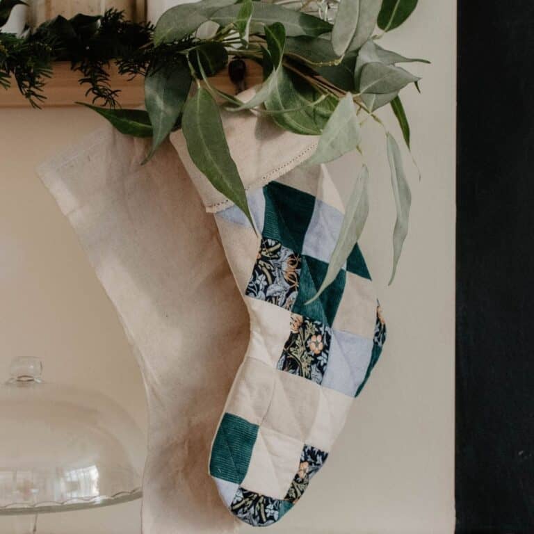 A patchwork stocking with green, blue, and patterned squares hangs beside a plain beige stocking. Both are adorned with green foliage at the top and dangle on a wall near a glass dome.