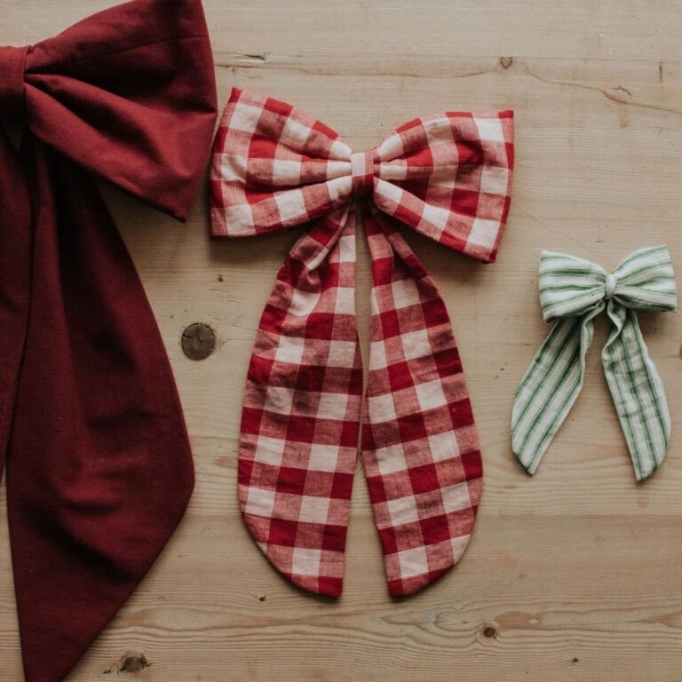 Three fabric bows on a wooden surface showcase how to make fabric bows: a large solid red bow on the left, a medium red and white checkered bow in the center, and a small green and white striped bow on the right.