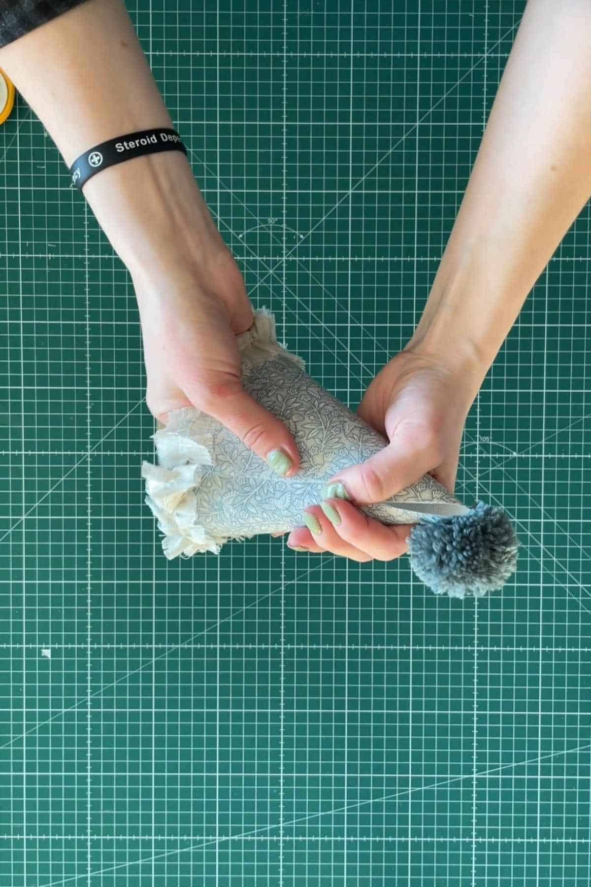 Hands holding a DIY party hat with a pom-pom on top, over a green cutting mat with a grid pattern.