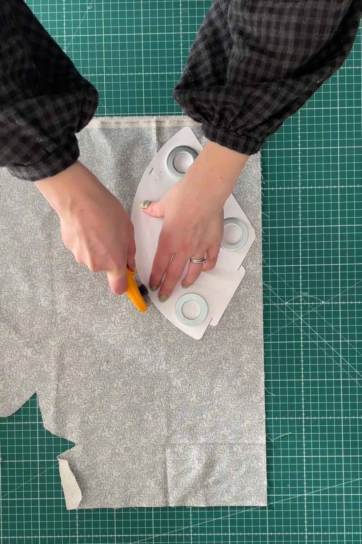 Person uses a rotary cutter to slice patterned fabric on a green grid cutting mat, following a free template secured with weights. 