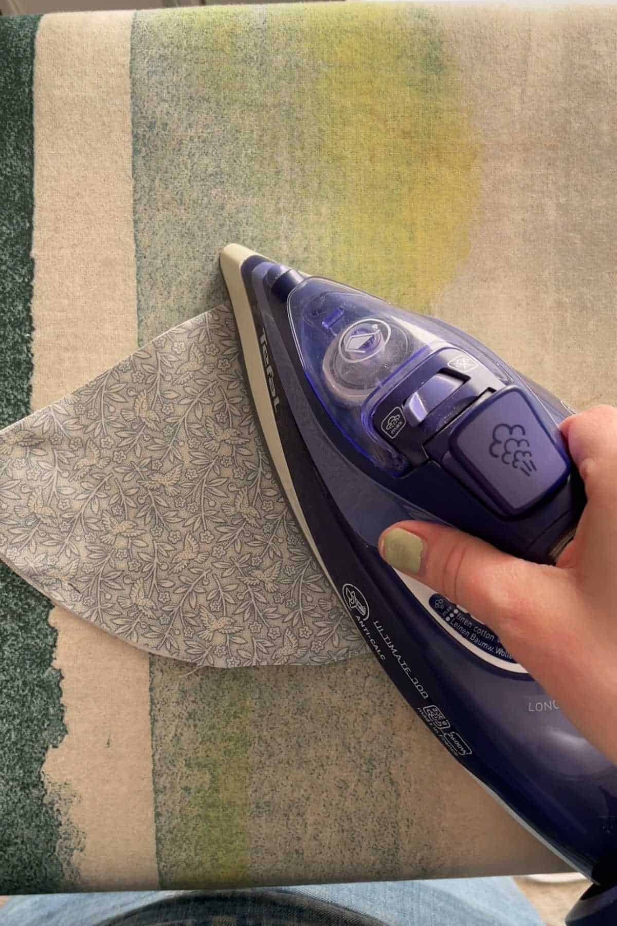A person is ironing a piece of fabric with a purple steam iron on a patterned ironing board cover. The fabric has a delicate floral design, and the persons fingernails are painted light green.