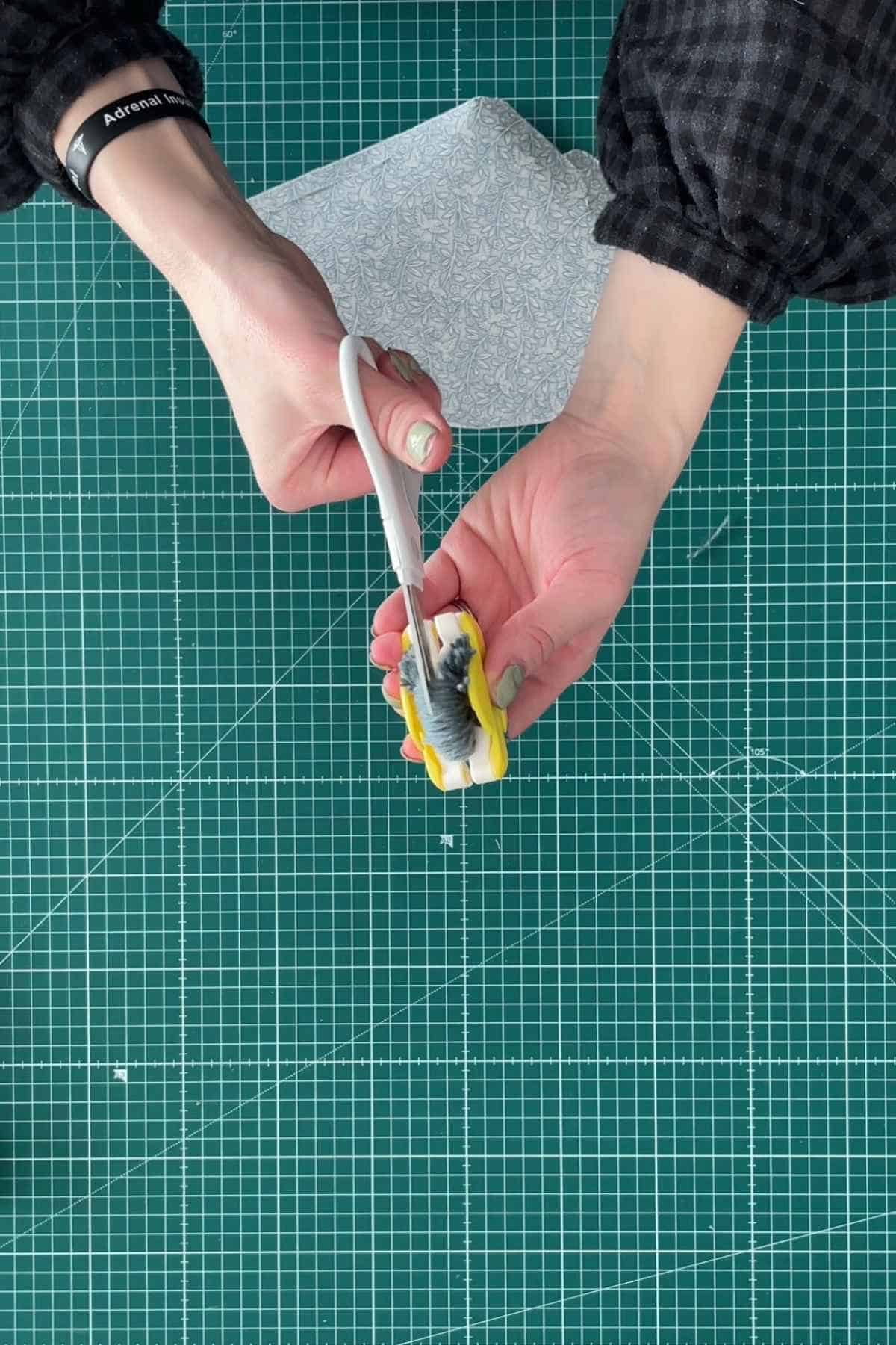 A person is making a pom pom in blue wool for the top of a DIY party hat. They work on a green cutting mat with grid lines. A fabric piece peeks from the top. 