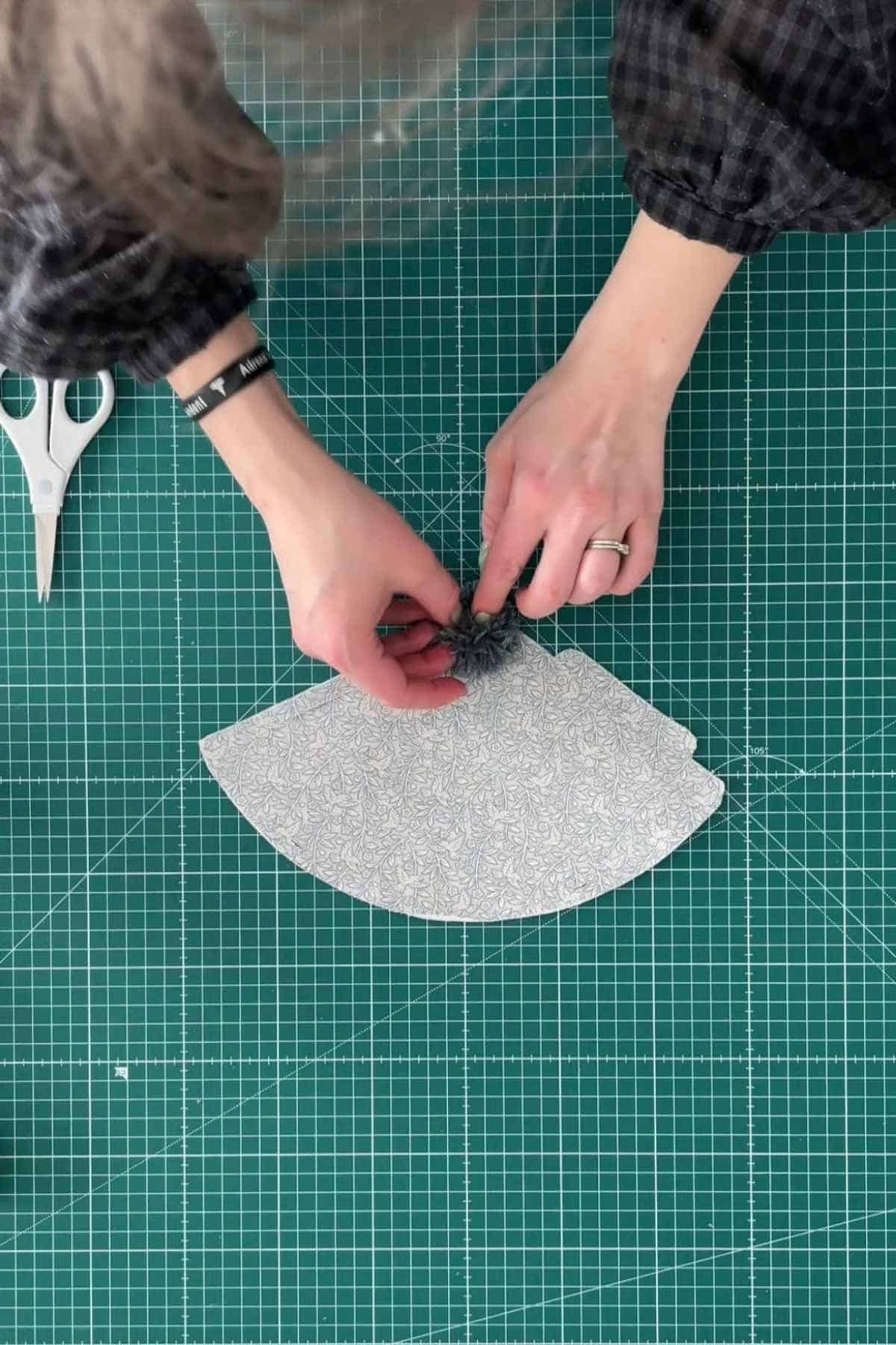 A person is crafting DIY party hats on a green cutting mat, applying the pom pom to the top of the hat. Scissors lay nearby.