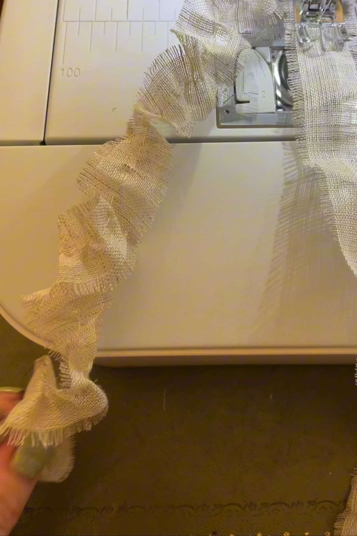A sewing machine stitches a length of light, ruffled fabric, creating the ruffled decorative edge for a DIY party hat. The fabric flows from the presser foot area as stitching progresses, while controls remain partially visible.