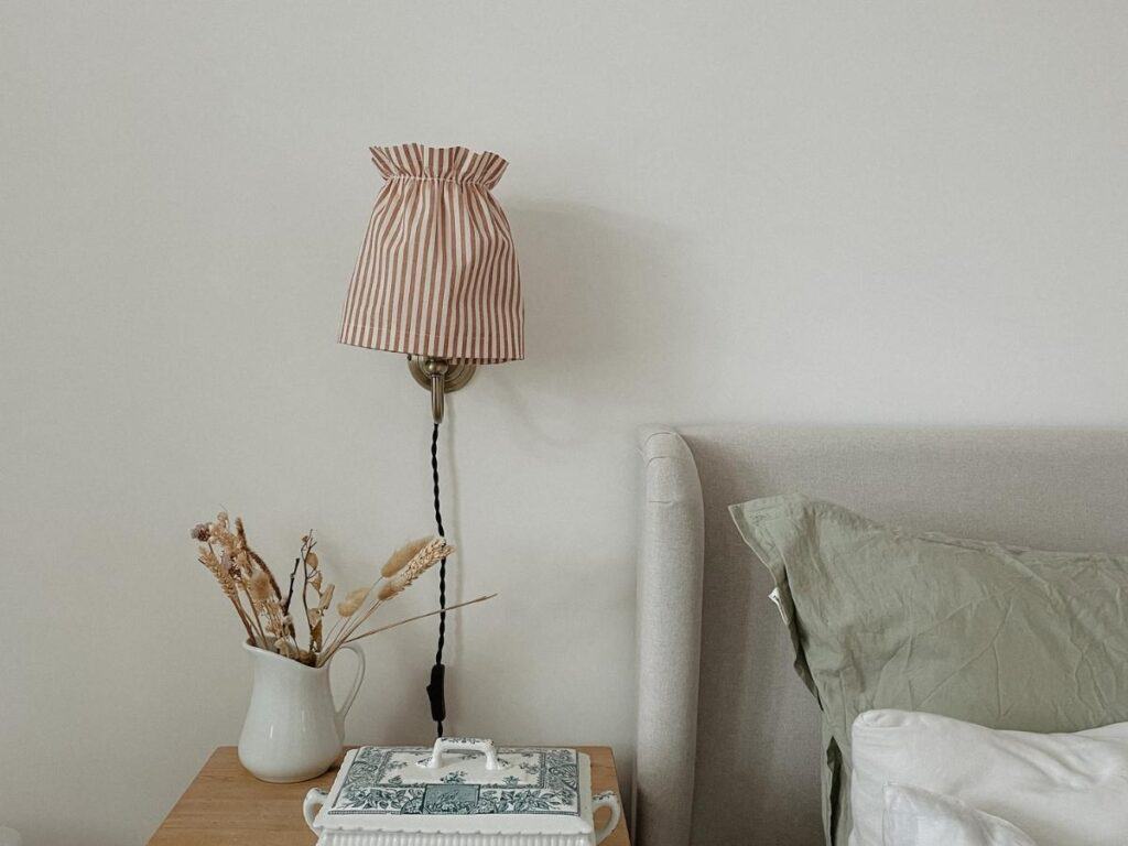 A red striped gathered lampshade cover on a wall lamp next to a grey linen bed. There is an antique pine bedside table next to the bed.