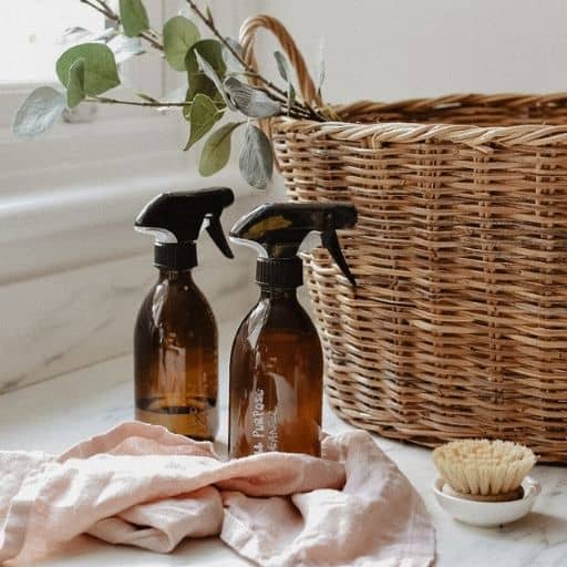Two amber spray bottles sit on a kitchen counter with a pink cloth and reusable washing up brush. In the background is a wicker laundry basket.