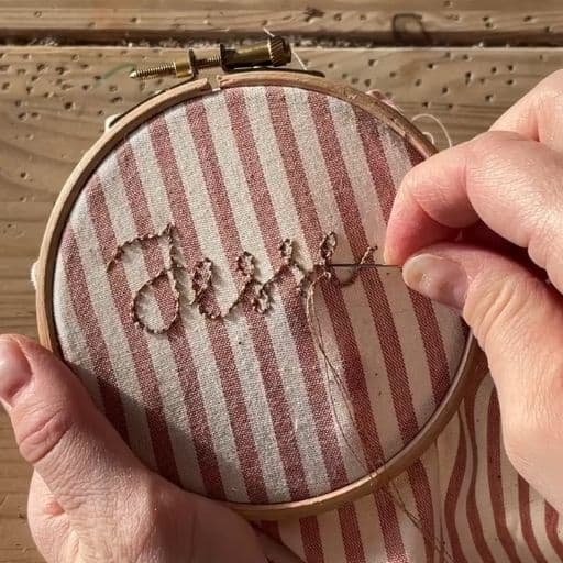 A woman embroiders the name Jesse in gold metallic thread on a red stripe vintage fabric