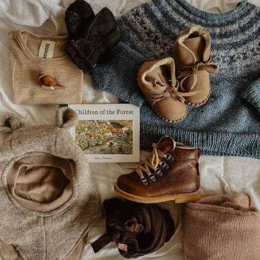 A flatlay showing a handknitted blue fairisle jumper, leather baby booties, merino wool top, balaclava, angulus boots and children of the forest book