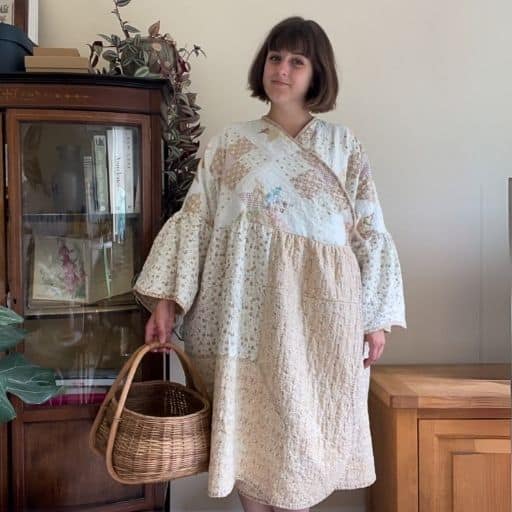 A woman stands in her cottage style living room wearing a handmade dress made from an antique quilt. She is holding a basket.