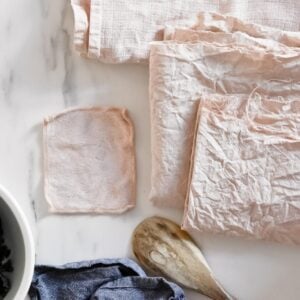 A collection of light pink squares of fabric, naturally dyed with avocado pits, is arranged on a white marble surface. A wooden spoon lies nearby, partially in view, next to a white-bottomed bowl. The fabric appears soft and gently wrinkled.