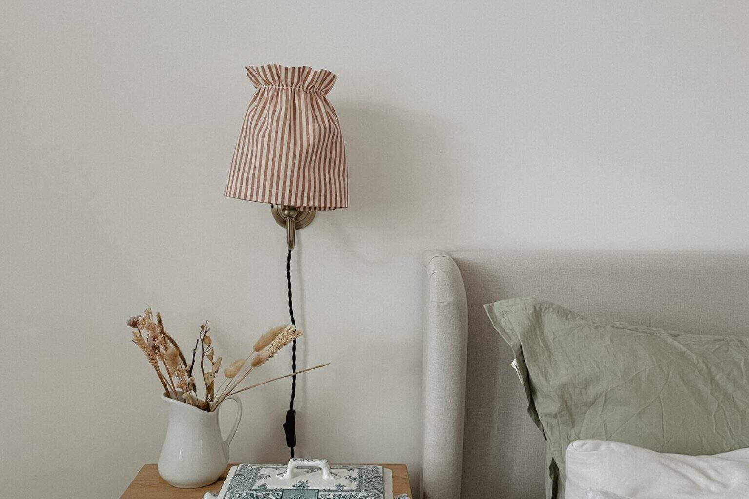 A bedside scene with a striped fabric lampshade on the wall above a nightstand. The nightstand holds a white jug with dried plants, a decorative book, and a tissue box. A pillow with a green pillowcase rests on a light gray bed.