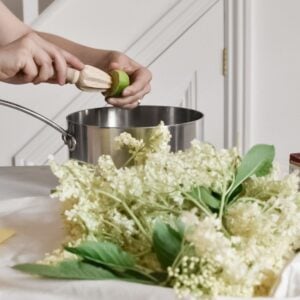 Hands using a wooden citrus reamer to juice a lime over a silver pot, crafting the perfect blend for elderflower cordial. In the foreground, freshly foraged white elderflowers with green leaves rest on the countertop. The background reveals a partial view of a white staircase.