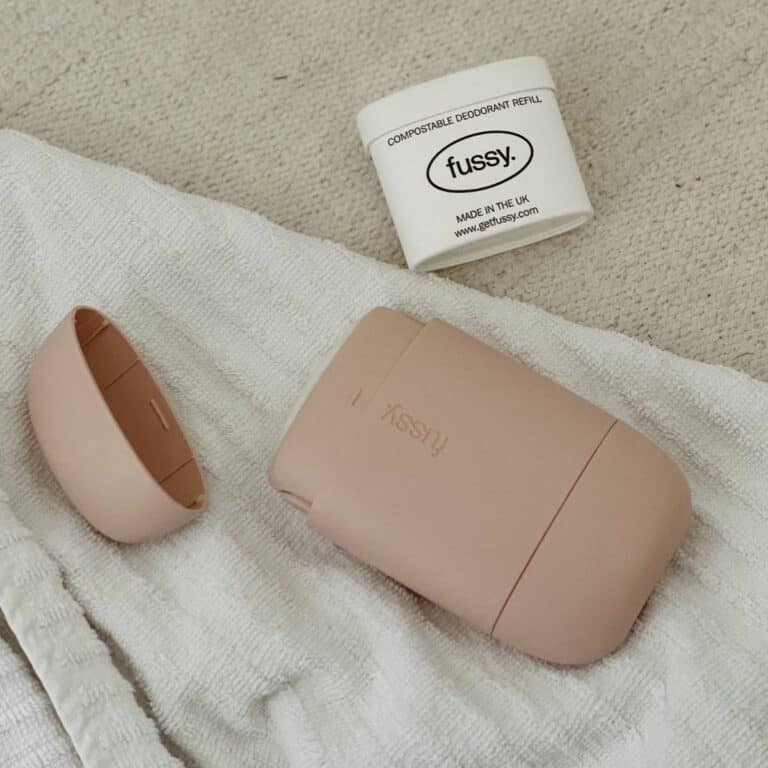 A beige fussy deodorant container and its cap are laid on a white towel. Next to them is a small white container labeled fussy, indicating it is a compostable deodorant refill made in the UK. The items are placed on a carpeted surface.