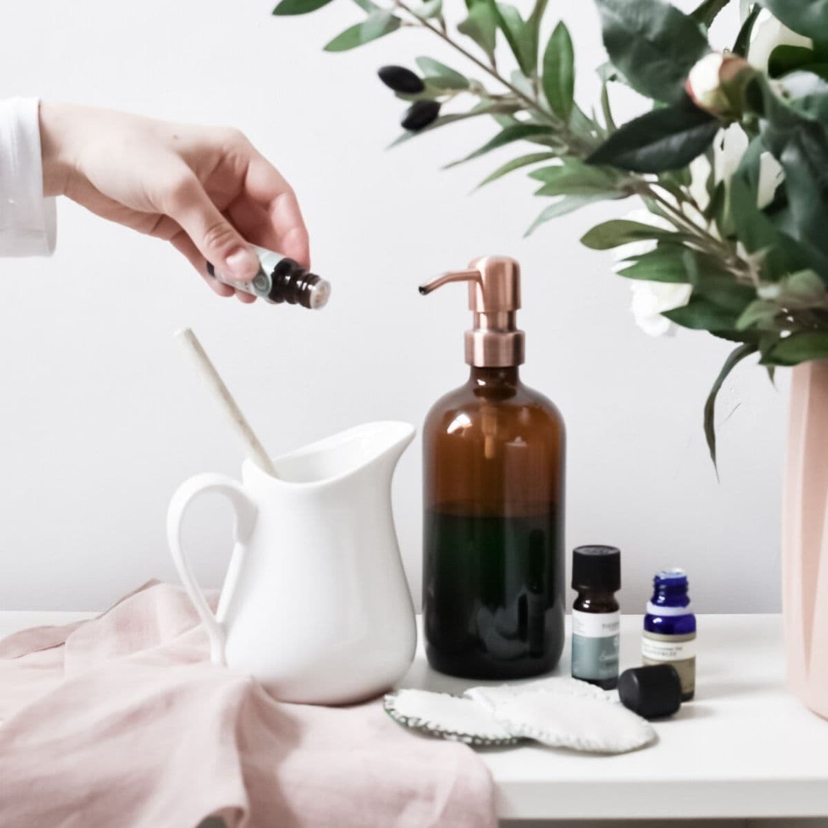 A hand delicately drops essential oil into a white pitcher on a pristine surface. Nearby, a brown glass soap dispenser filled with Castile soap hints at creating a natural homemade face-wash, surrounded by bottles of essential oils, a soft pink cloth, knitted pad, and leafy plant.