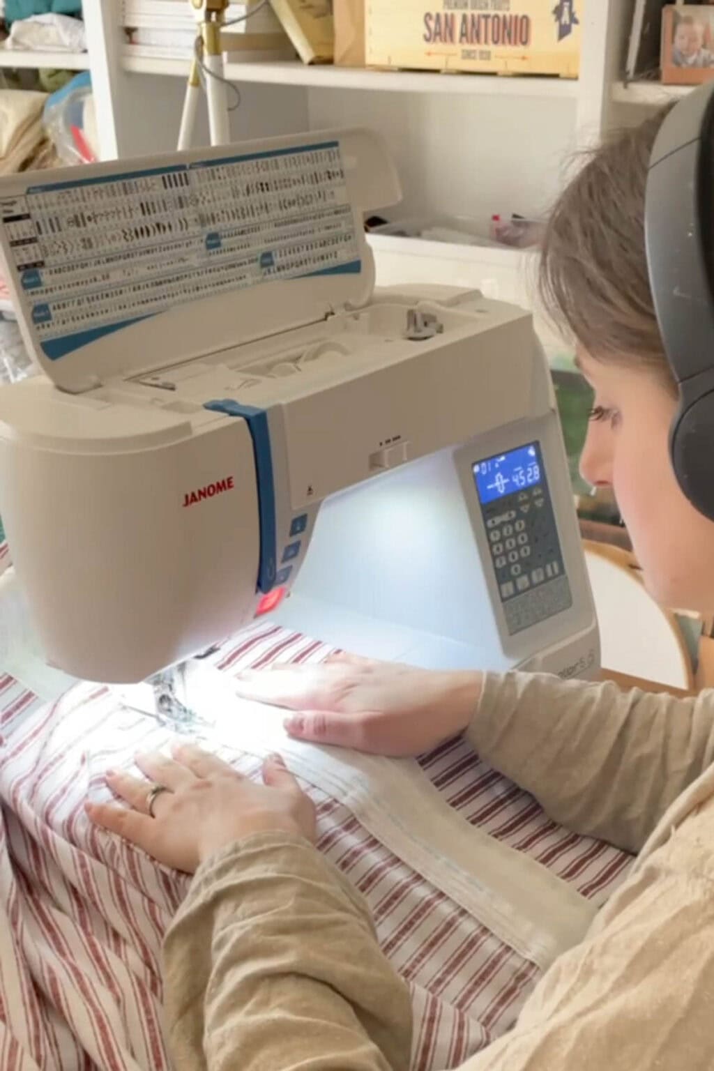 A person wearing headphones is focused on sewing a striped fabric, perhaps hinting at how to make curtains, using a Janome sewing machine. In the background, shelves filled with various sewing materials provide ample inspiration for creations.