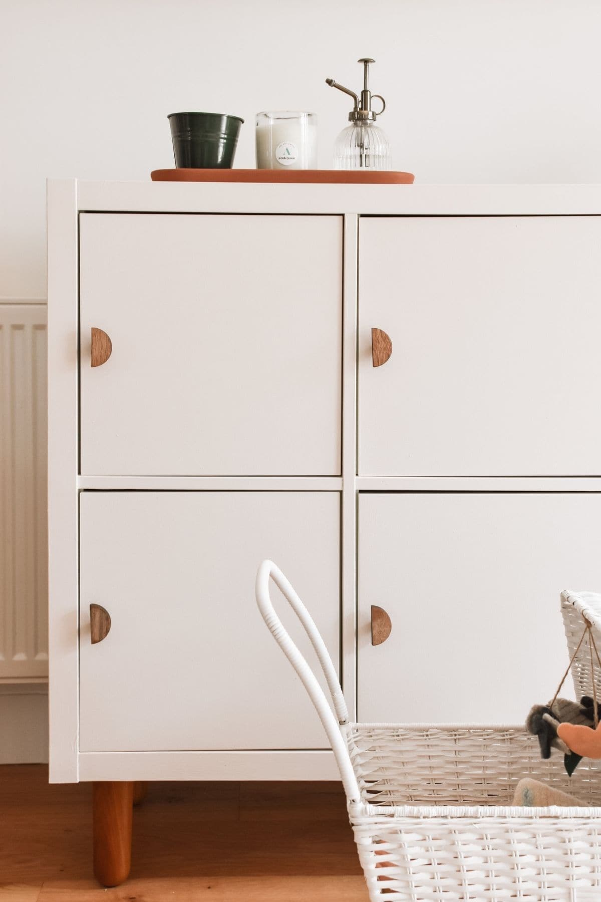 Minimalist room corner showcasing a clever IKEA Kallax hack, with a pale pink cabinet and wooden handles . Topped with a green pot, candle, and glass mister, its paired with a white wicker toy pushchair on the wooden floor.