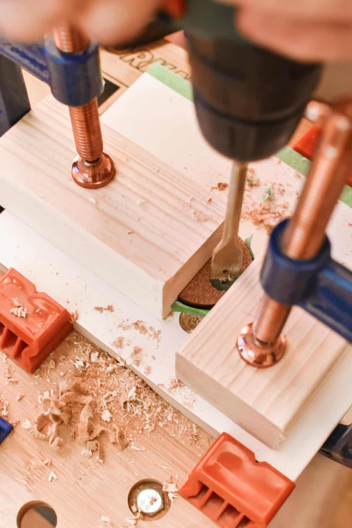 A close-up of a woodworking setup shows a drill making precise holes in a plywood shape. Two clamps keep the plank sturdy as wood shavings scatter around, while a green ruler rests in the background.