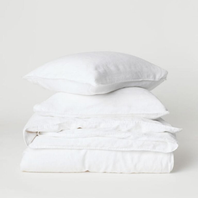 A neatly folded stack of fresh white linens, including two pillows and sheets, against a plain background.