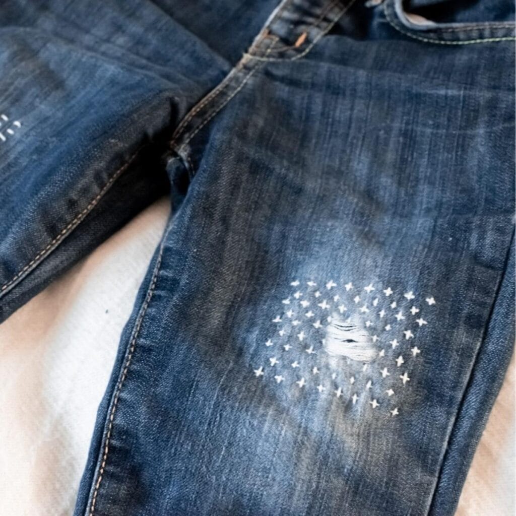 A pair of blue jeans featuring a white embroidered sashiko patch with small crosses around a distressed hole on one leg, laid out on a light surface.