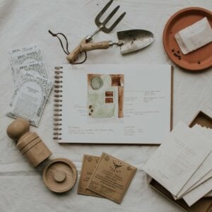 A flat lay of garden planning items on a white cloth reveals plans for a small cottage garden. Includes a sketchbook with a 2023 design, seed packets, a trowel, a gardening fork, and wooden containers. Theres also a small plate with seeds and a piece of paper.