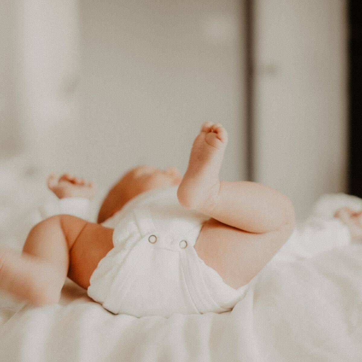A newborn baby wearing a white nappy lies on a soft, white bed with their feet playfully in the air. The room is softly lit, creating a cozy and warm atmosphere.