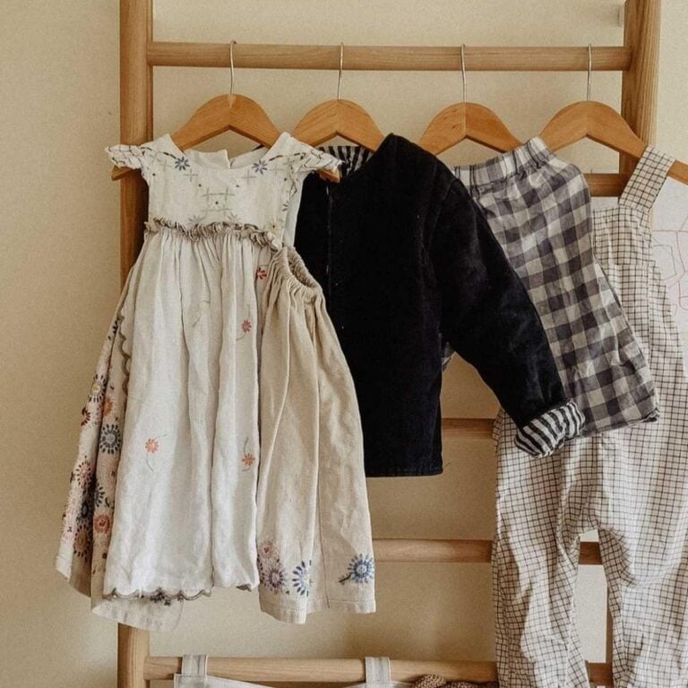 Three outfits made from childrens sewing patterns hang on wooden hangers: a floral embroidered dress, a black jacket with checked pants, and light grid-patterned overalls, all displayed on a wooden rack against a beige wall.