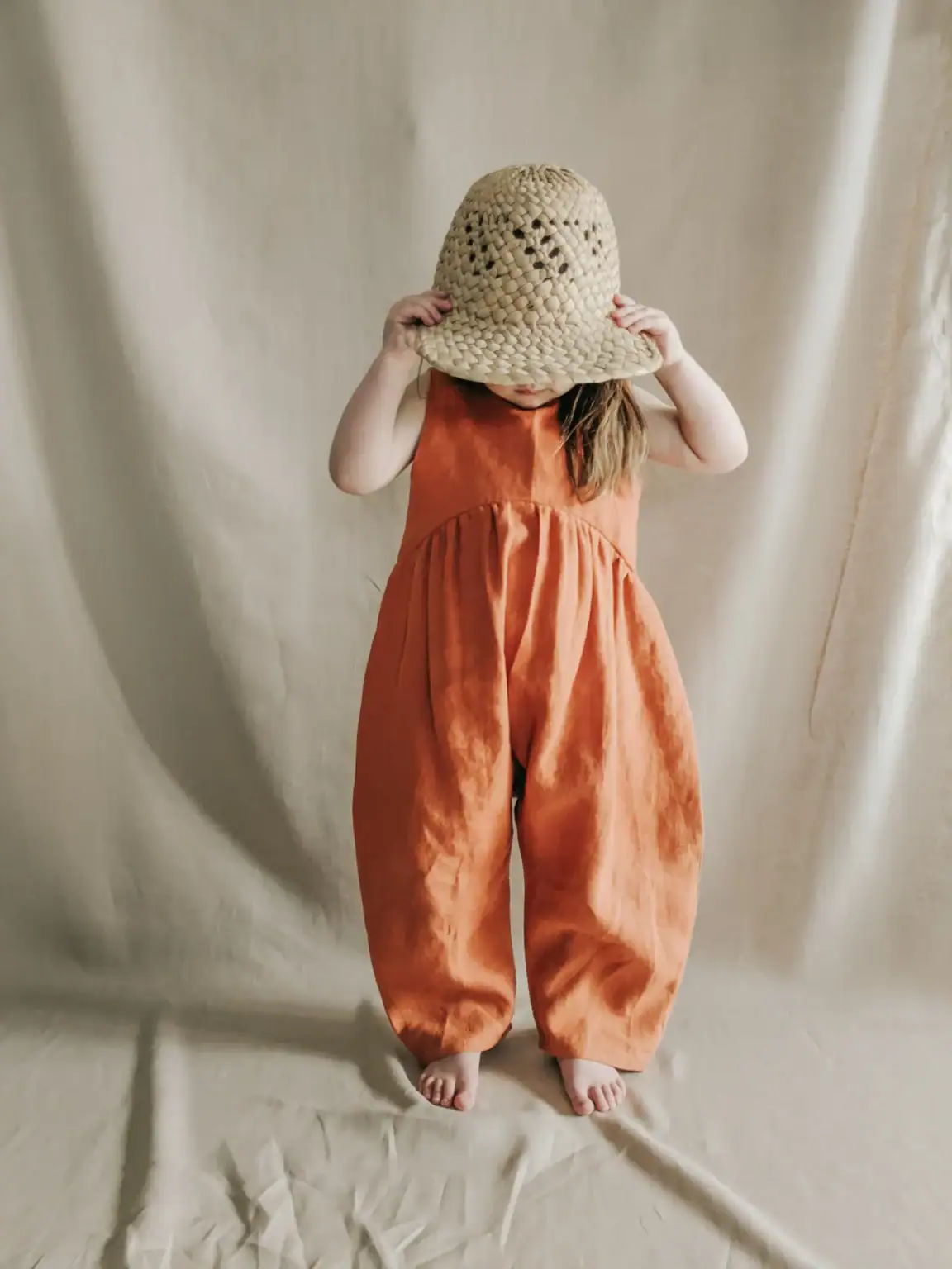 A young child in a loose orange jumpsuit stands barefoot against a neutral backdrop, holding a woven straw hat over their face and obscuring their features.