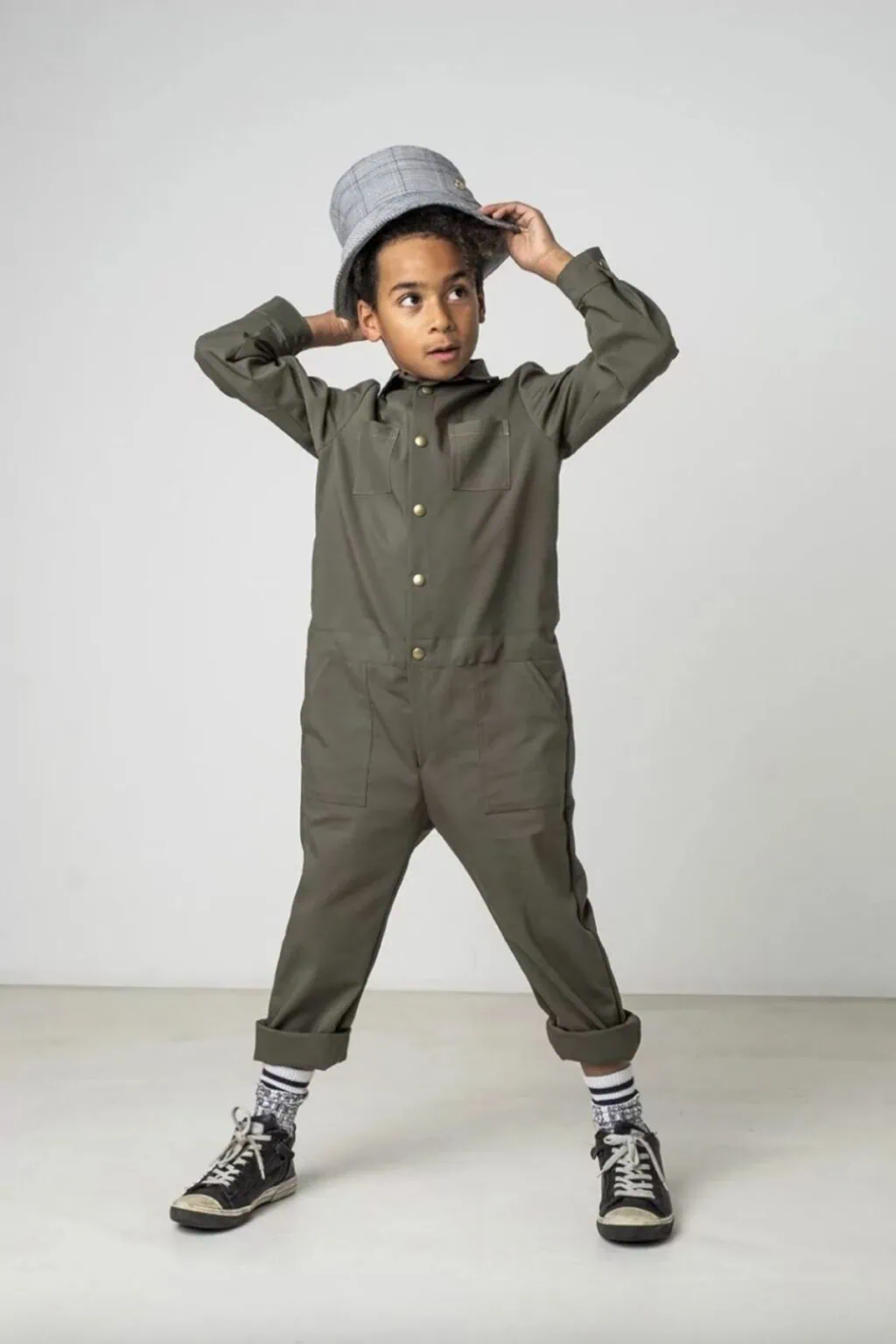 A child in a green jumpsuit madew with the Indigo jumpsuit sewing pattern and black high-top sneakers stands with legs apart, holding a gray plaid hat on their head, looking to the side against a plain light background.