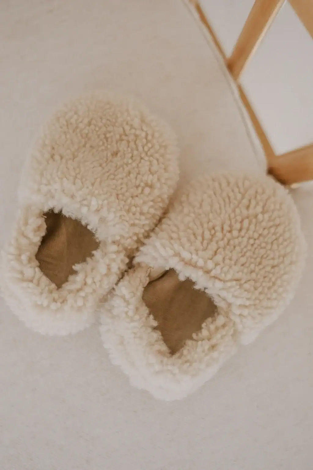 A pair of cozy, cream-colored, fluffy slippers with soft, textured fabric sits on a light surface near a wooden chair leg.