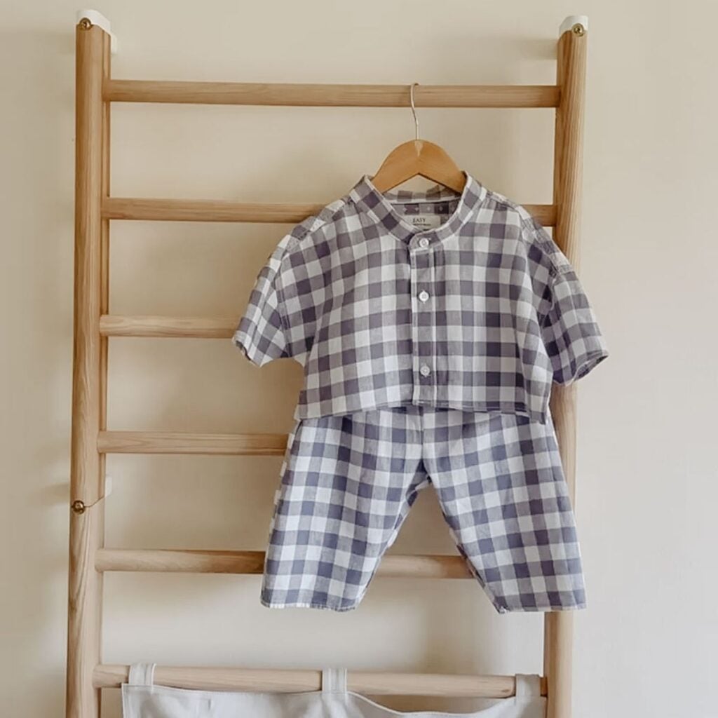 An upcycled baby outfit in baby blue and white checks, with short sleeves and buttons, hangs on a wooden ladder against a beige wall. A fabric organizer is attached to the lower part of the ladder.