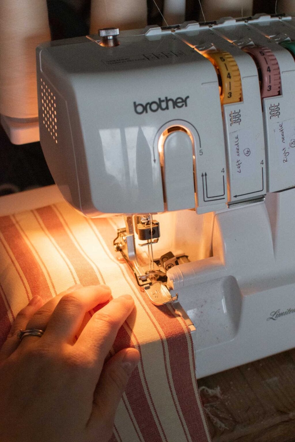 A close-up of a person’s hand guiding striped fabric through a Brother sewing machine, illuminated by a built-in light—perfect for crafting DIY curtains to display in your glass cabinet.