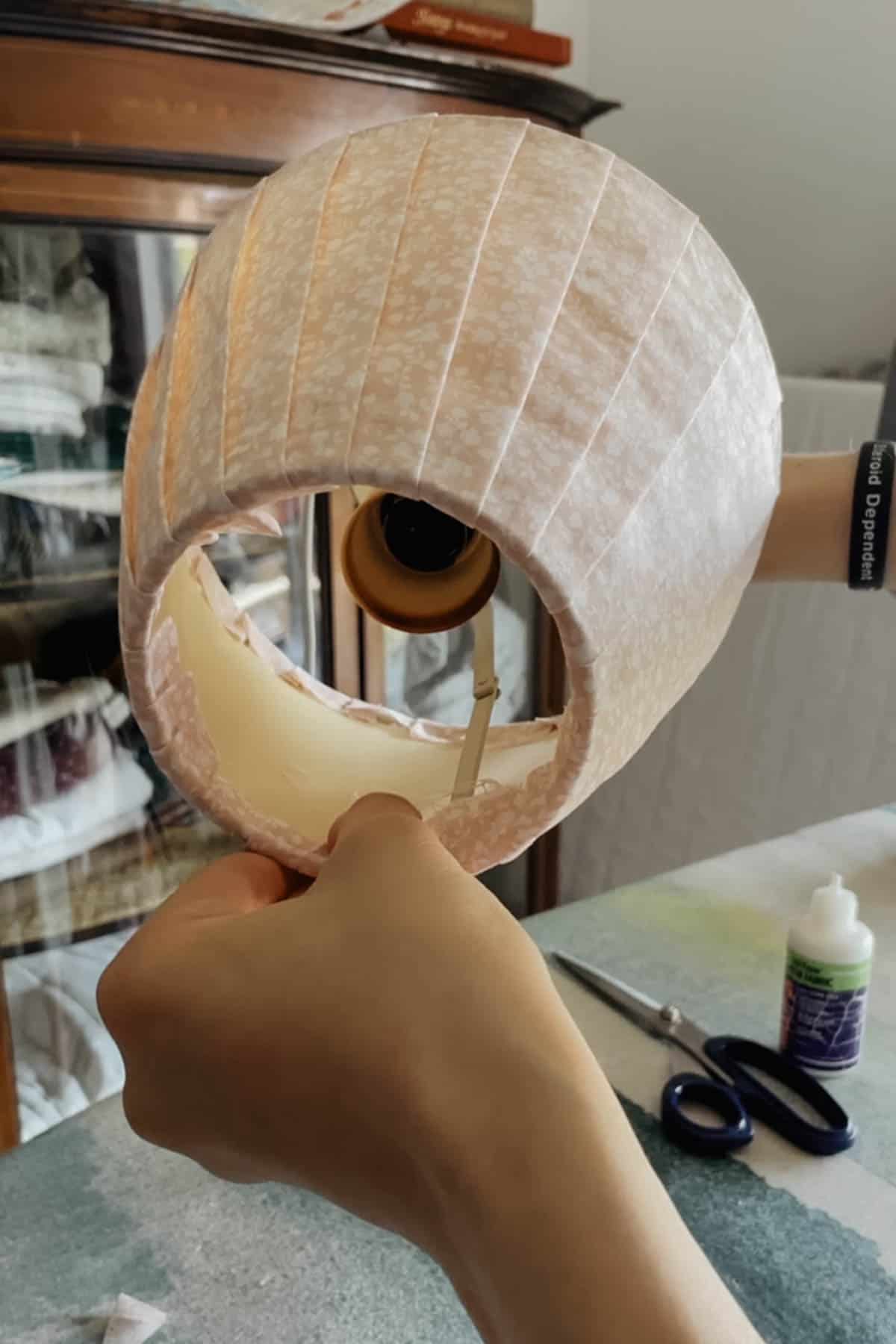 A person is pressing the raw edges of some fabric strips onto the inside of the lampshade