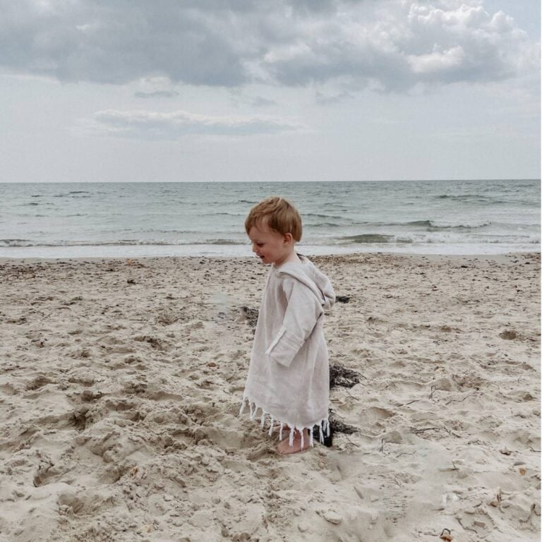 A little boy in a beige hooded towel poncho standing on the beach