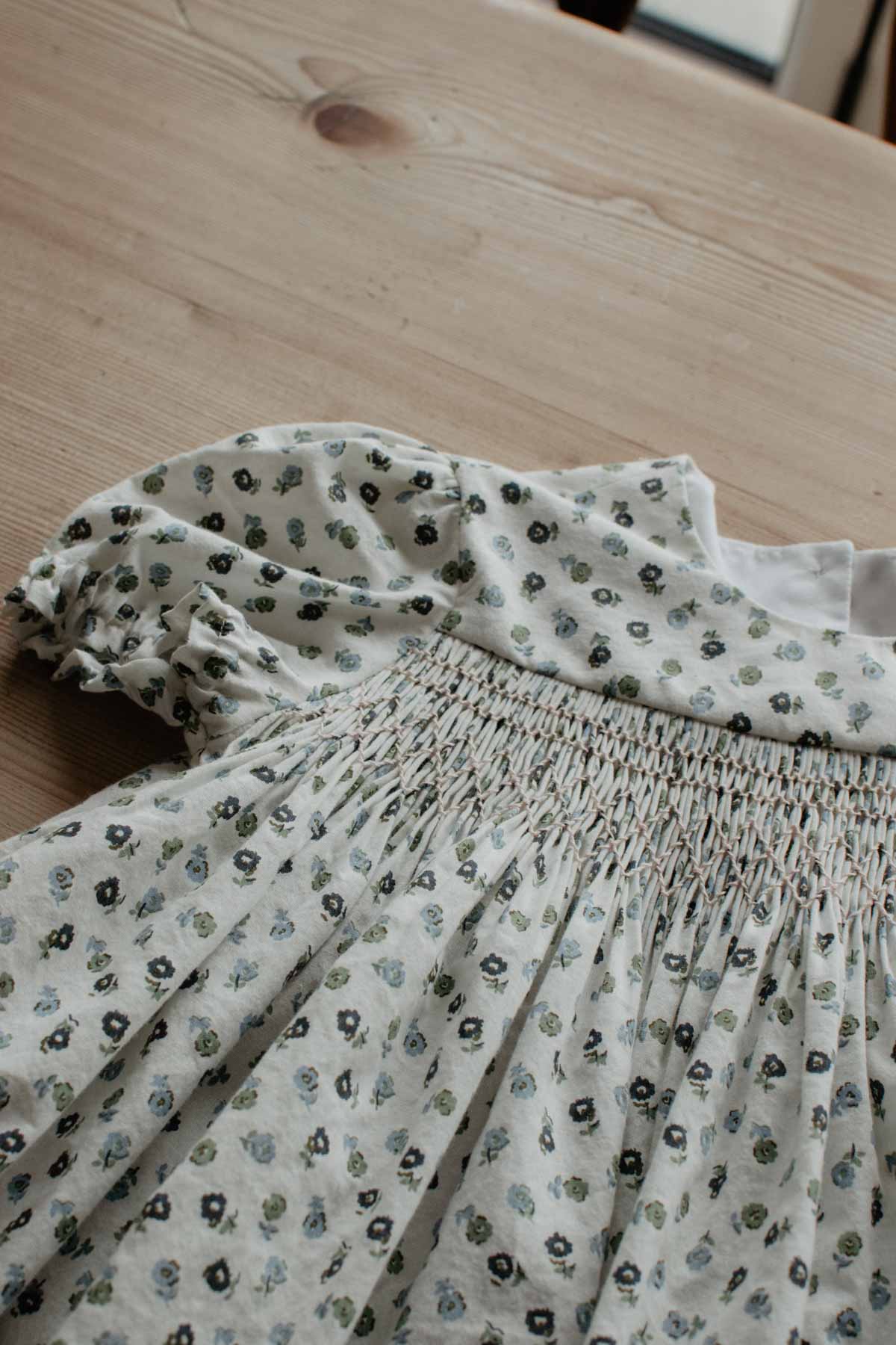 A little handmade smocked dress lies on a wooden table.