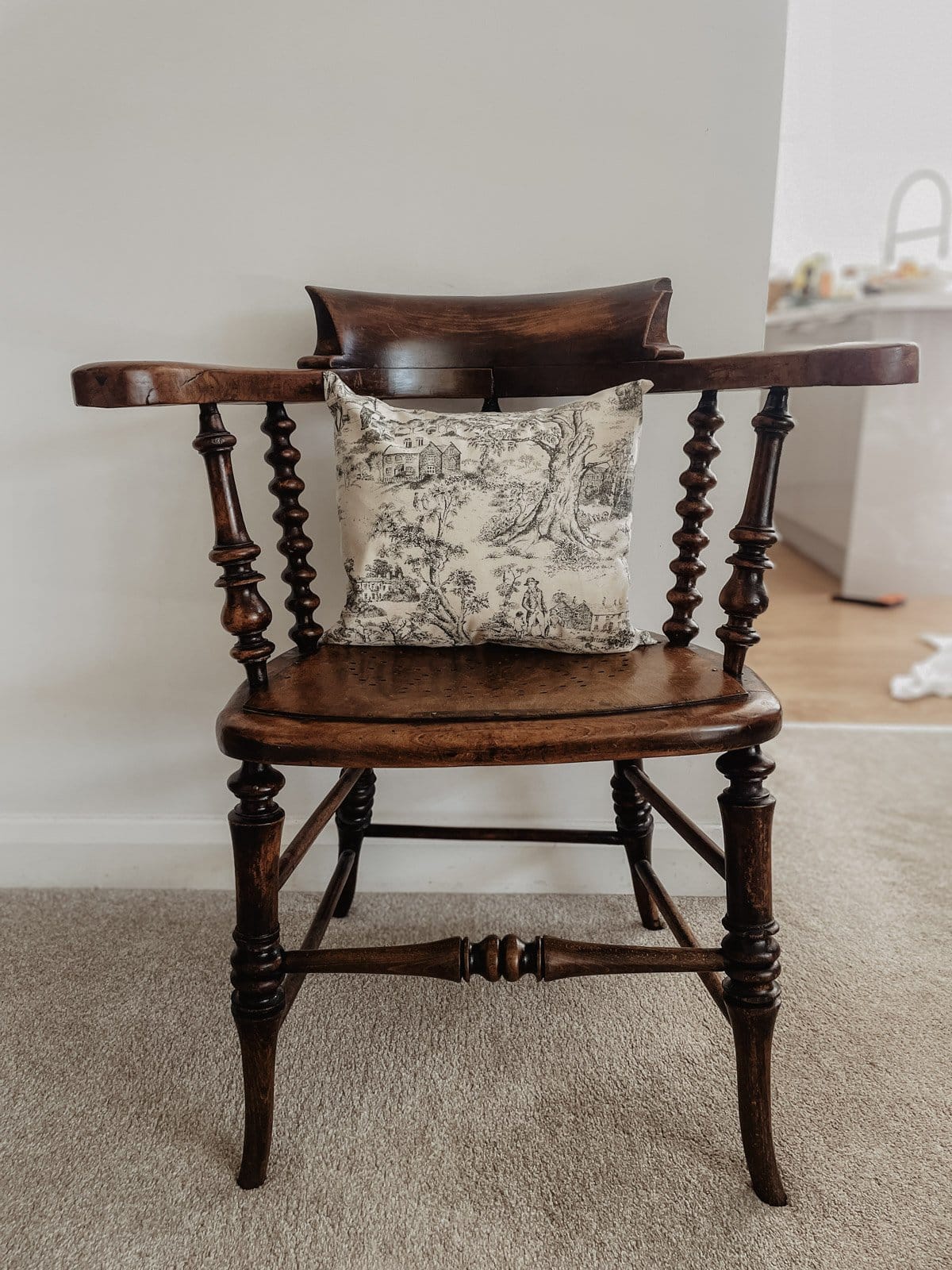 A toile print pillow sits on an antique wooden captain's chair. There is laundry on the floor and dishes in the background.
