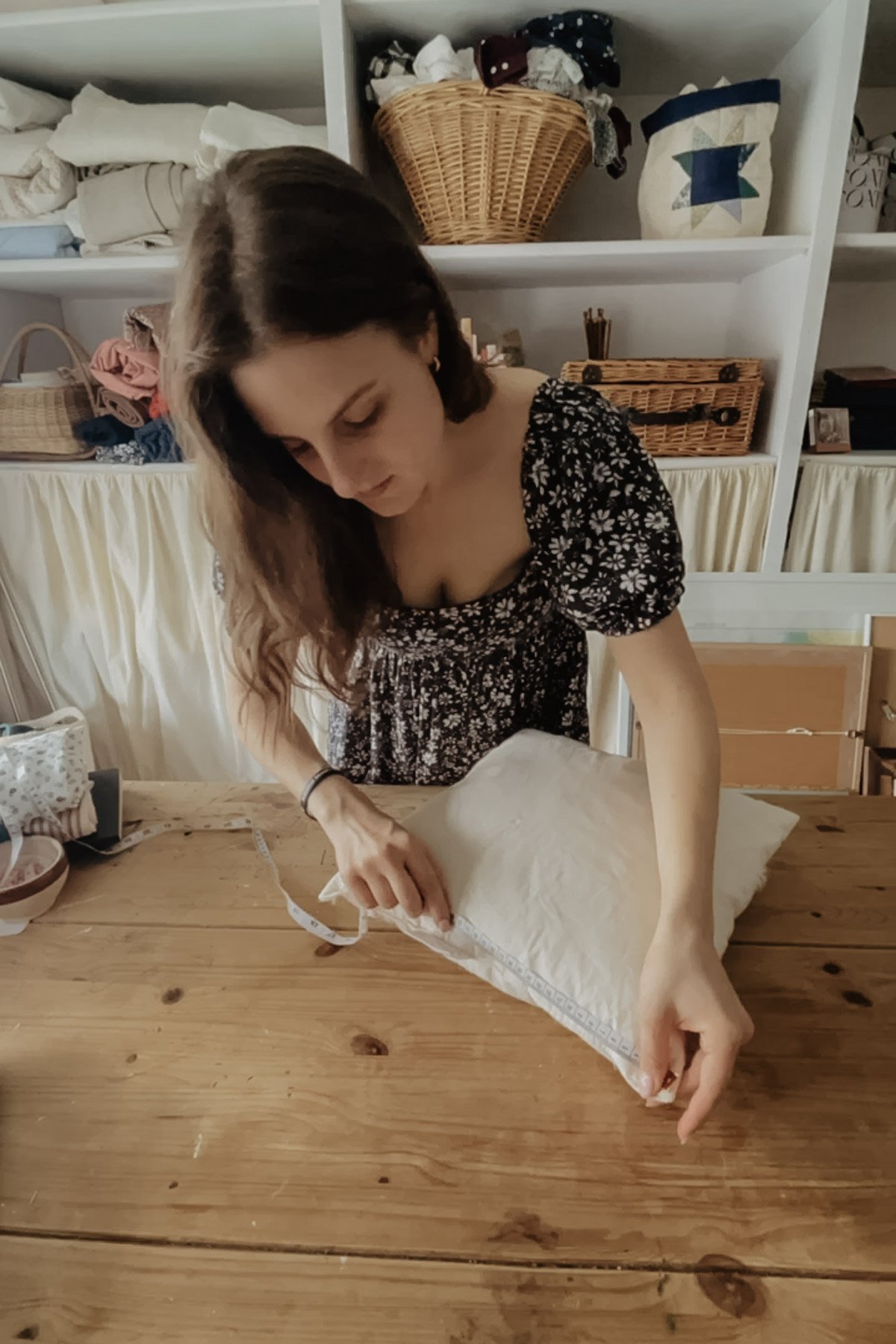 A woman measures the height of a pillow insert using a tape measure. There are baskets and piles of fabric in the background.