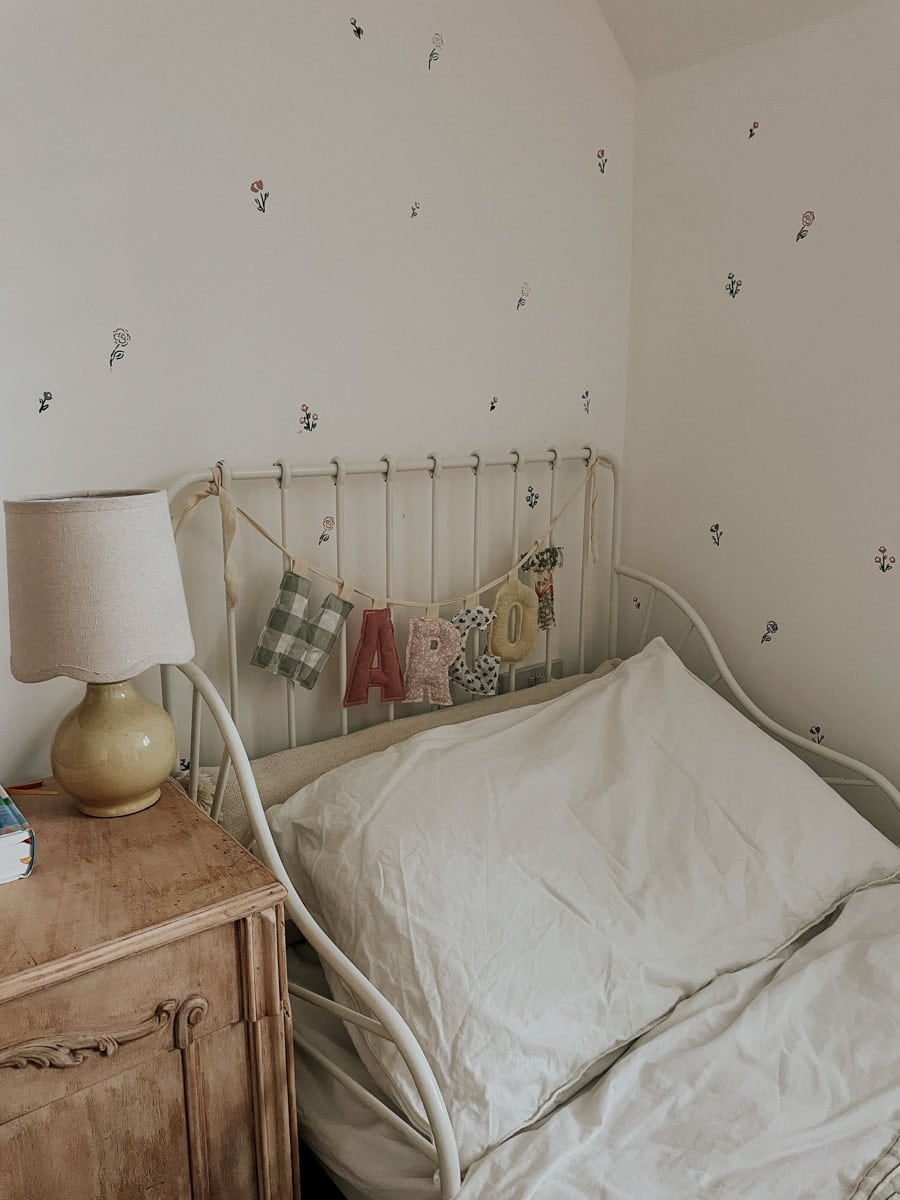 A hand stencilled wall behind a bed with a banner reading 'MARGOT' and a yellow lamp