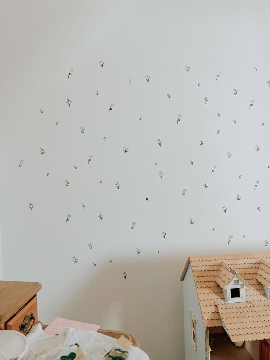 A wall with hand stencilled flowers in pink and blue. A dollhouse is in the bottom right hand corner.
