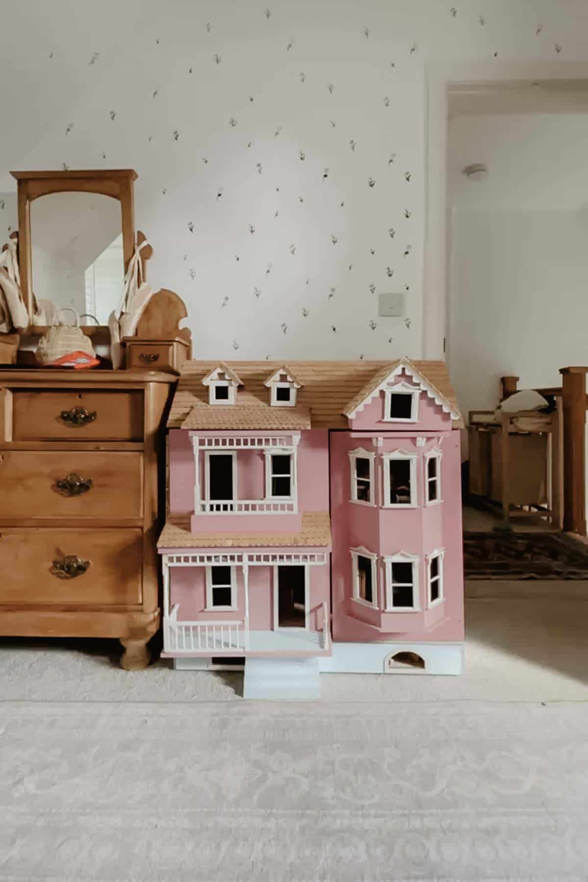 A pink dollhouse with white windows and doors. It sits next to an antique dresser.