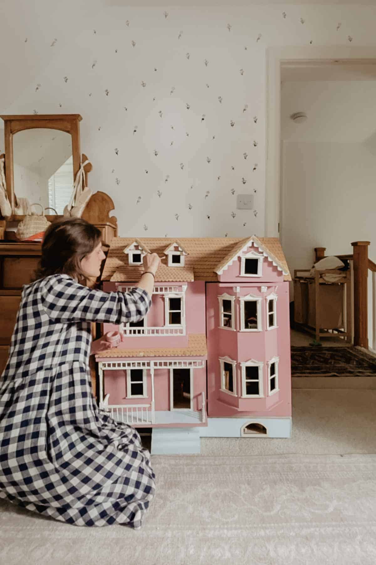 A lady paints a dollhouse with a muted pink paint. Floral walls and an antique dresser are in the background