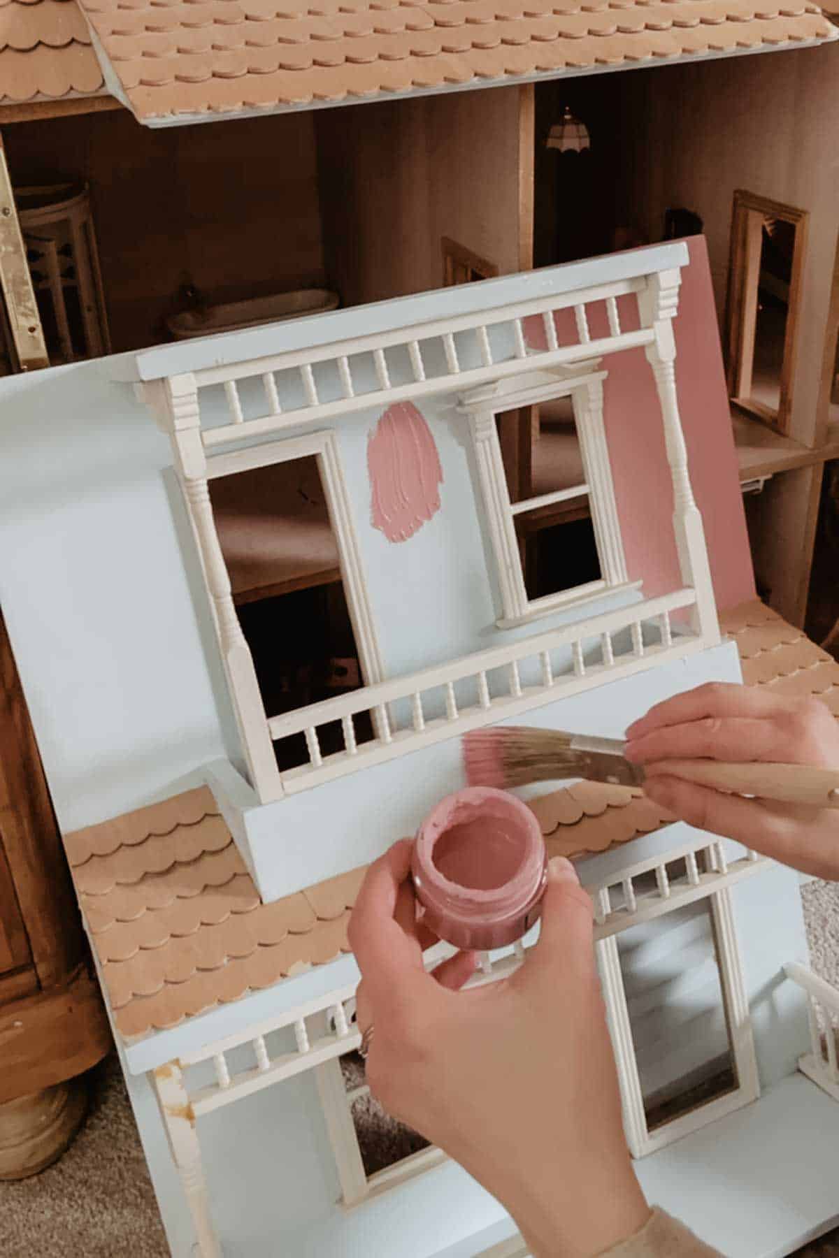 A person paints a dollhouse with a muted pink paint and small brush. 